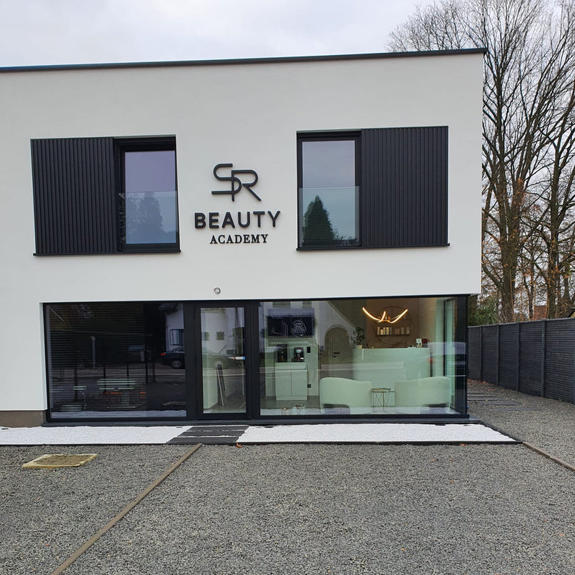 The exterior of a modern white and black building with large glass windows, featuring the text 'SR BEAUTY ACADEMY' on the facade. The ground floor has a glass-front reception area with white furniture inside, and the gravel driveway is in the foreground.