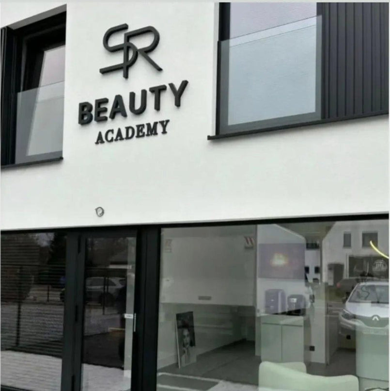 The exterior of a modern building facade featuring a white wall with black 3D lettering that reads 'SR BEAUTY ACADEMY' above a glass entrance door.